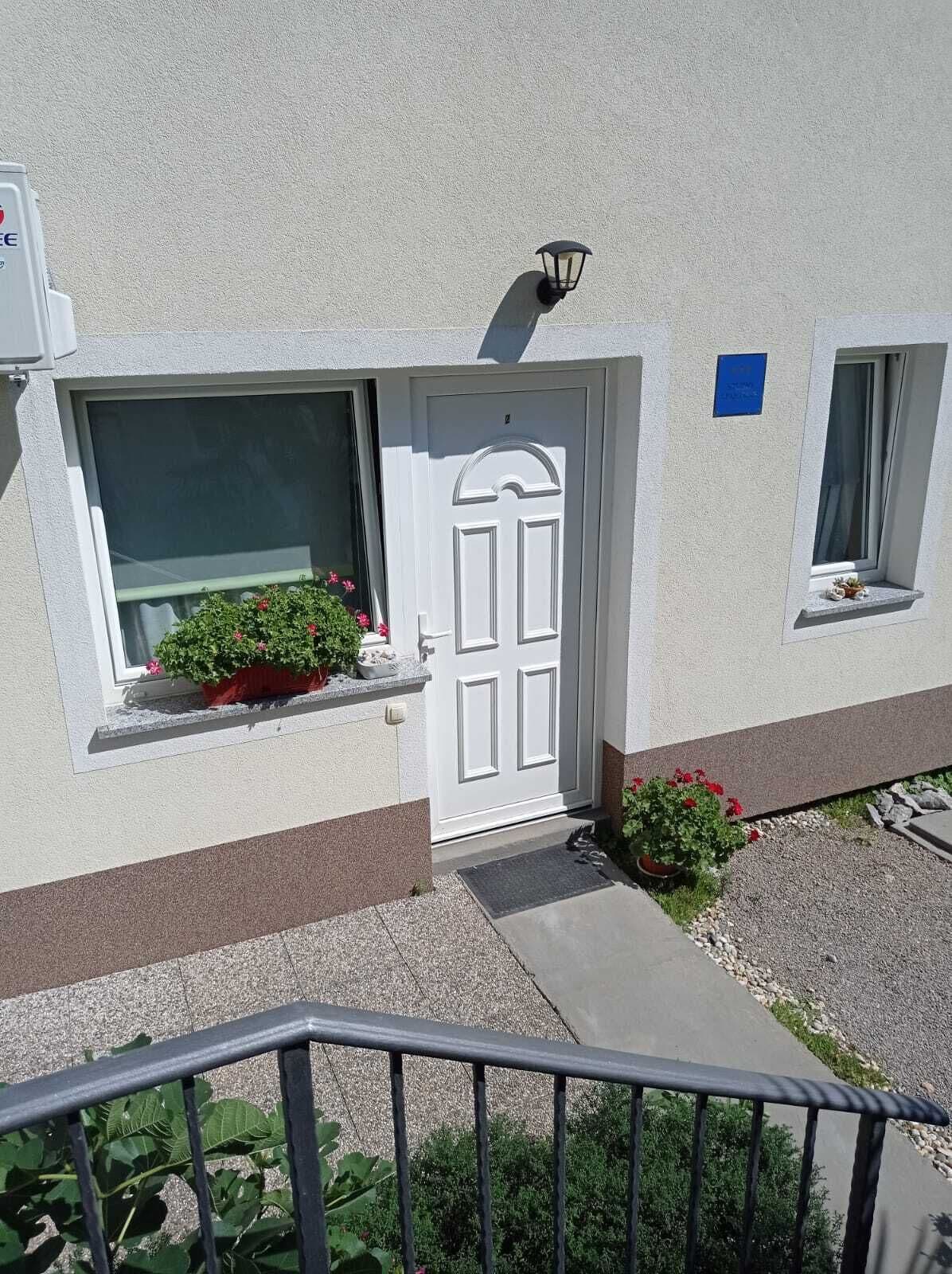 Un bâtiment avec une porte blanche, une fenêtre ornée d’un pot de fleurs et une petite fenêtre avec un rideau.