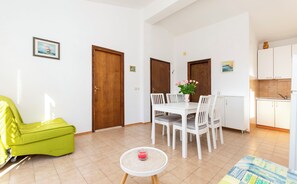 Un soggiorno luminoso con una poltrona verde, un tavolo da pranzo con sedie, un piccolo tavolino con una candela e un angolo cottura con pensili e lavello.
