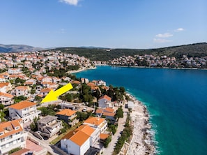 Une ville côtière aux bâtiments aux toits de tuiles rouges, bordée par une mer d’un bleu limpide, et où une flèche jaune indique un endroit précis.