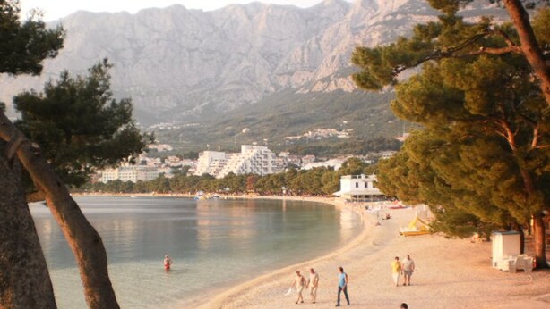 A beach with people walking and sitting, a clear sky, and mountains in the background. - Apartments Mise (Makarska)