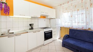 A modern kitchen with white cabinets, a stove, and a sink. There is a blue sofa and a window with curtains. - Apartments Mise (Makarska)