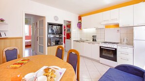 A modern kitchen with white cabinets, a stove, and a refrigerator. There is a dining table with chairs and a breakfast tray. - Apartments Mise (Makarska)