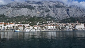A coastal town with buildings along the waterfront, boats docked, and mountains in the background. - Apartments Mise (Makarska)