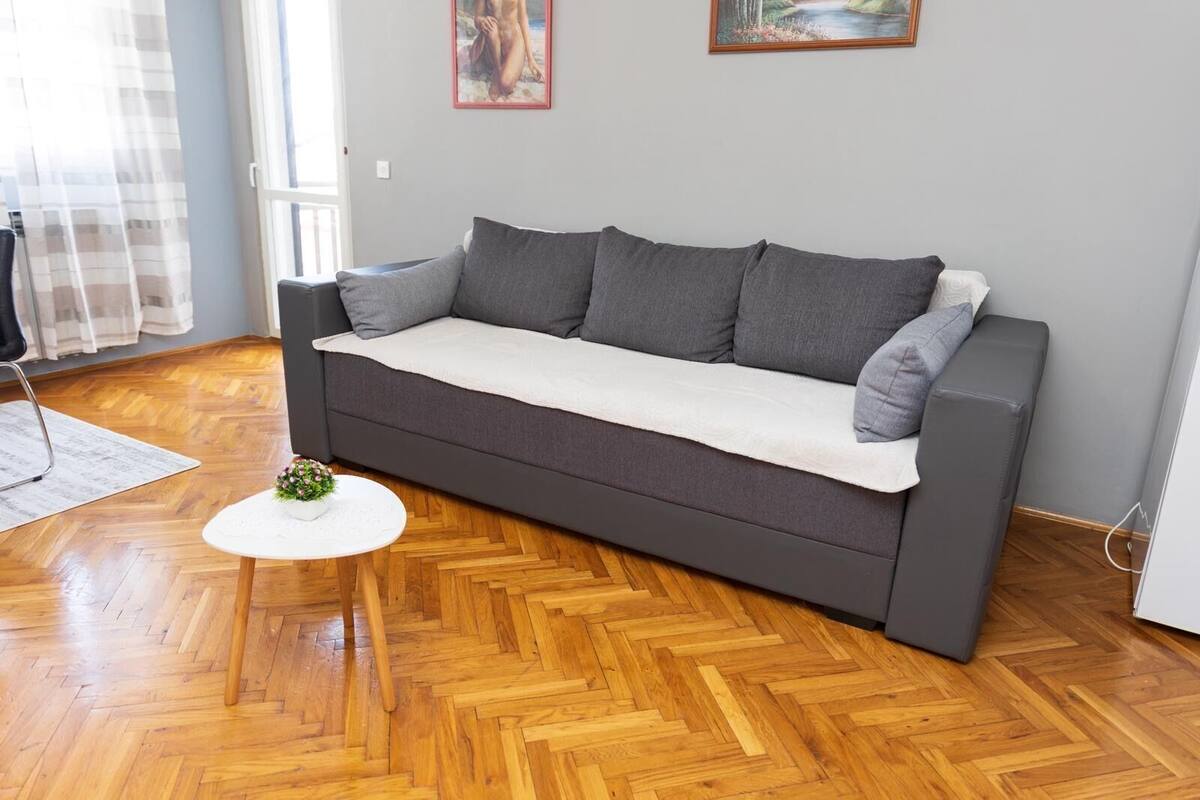 Un canapé gris surmonté d’un plaid blanc, une petite table blanche avec une plante, et un parquet en bois disposé en chevrons.