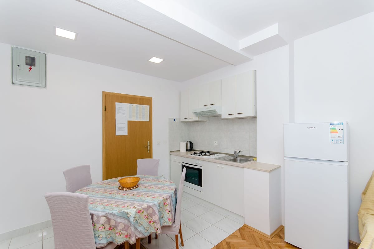 Une petite cuisine propre, avec des meubles blancs, un réfrigérateur, une cuisinière et un évier. On y trouve une table à manger avec des chaises et un panier posé dessus.