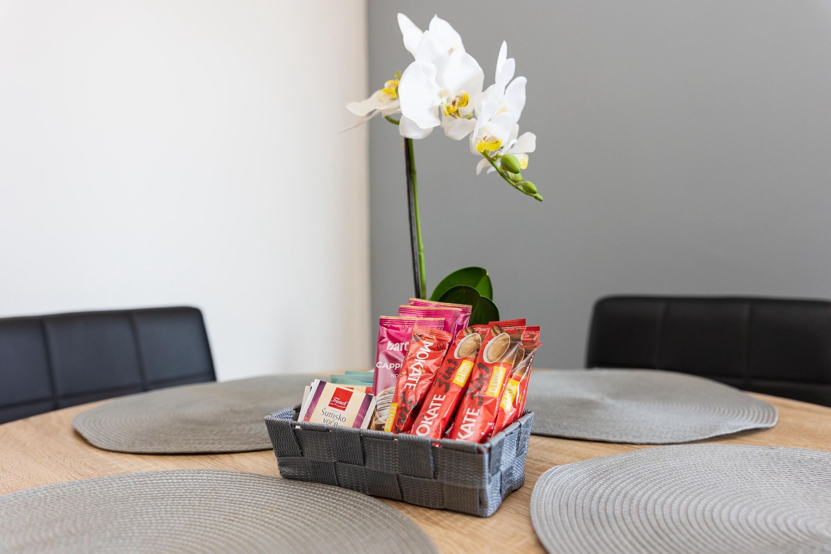 Une table à manger avec un panier de collations emballées, une orchidée blanche et des sets de table gris.