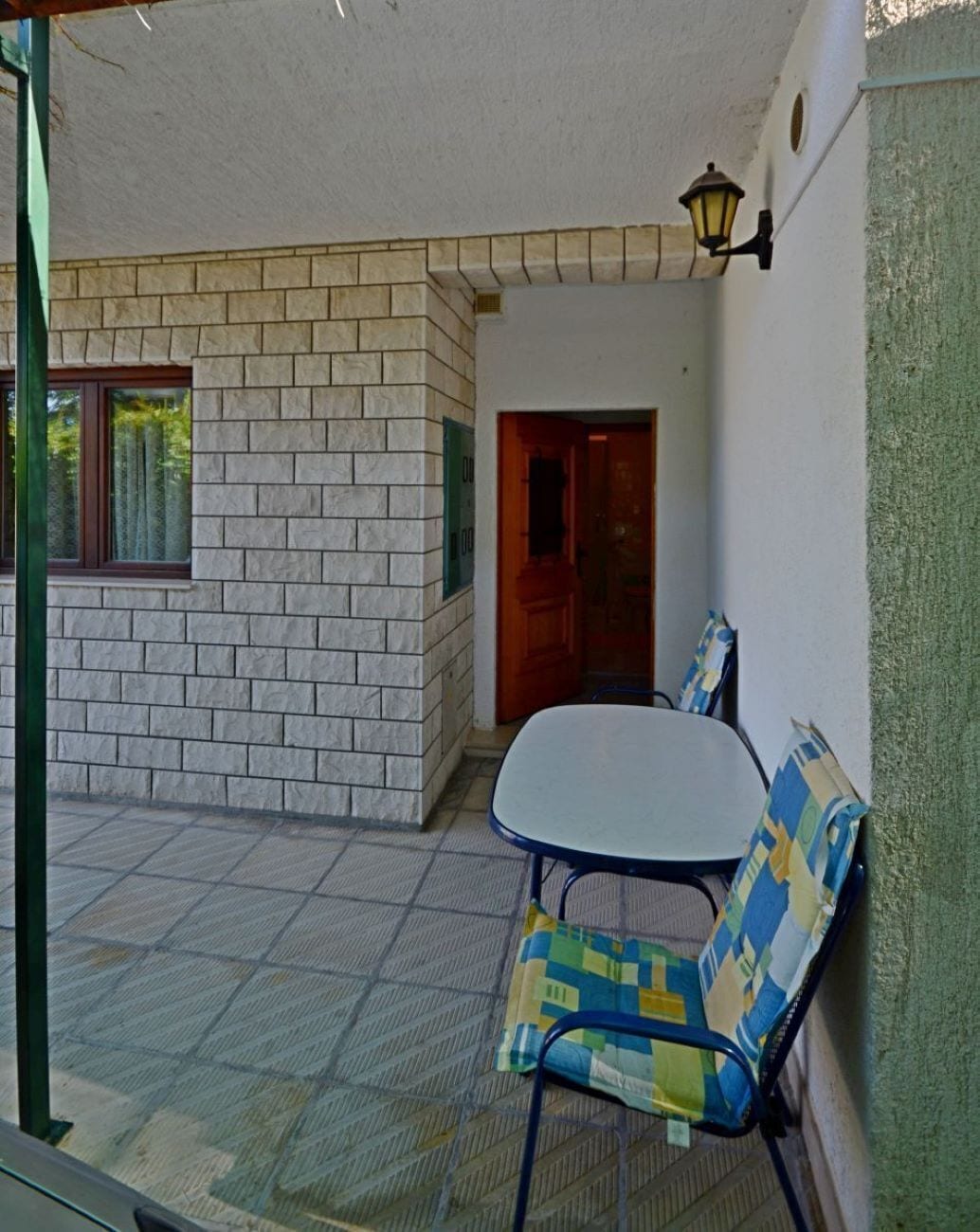 A covered patio with a table and chairs, a door, and a wall-mounted lamp.