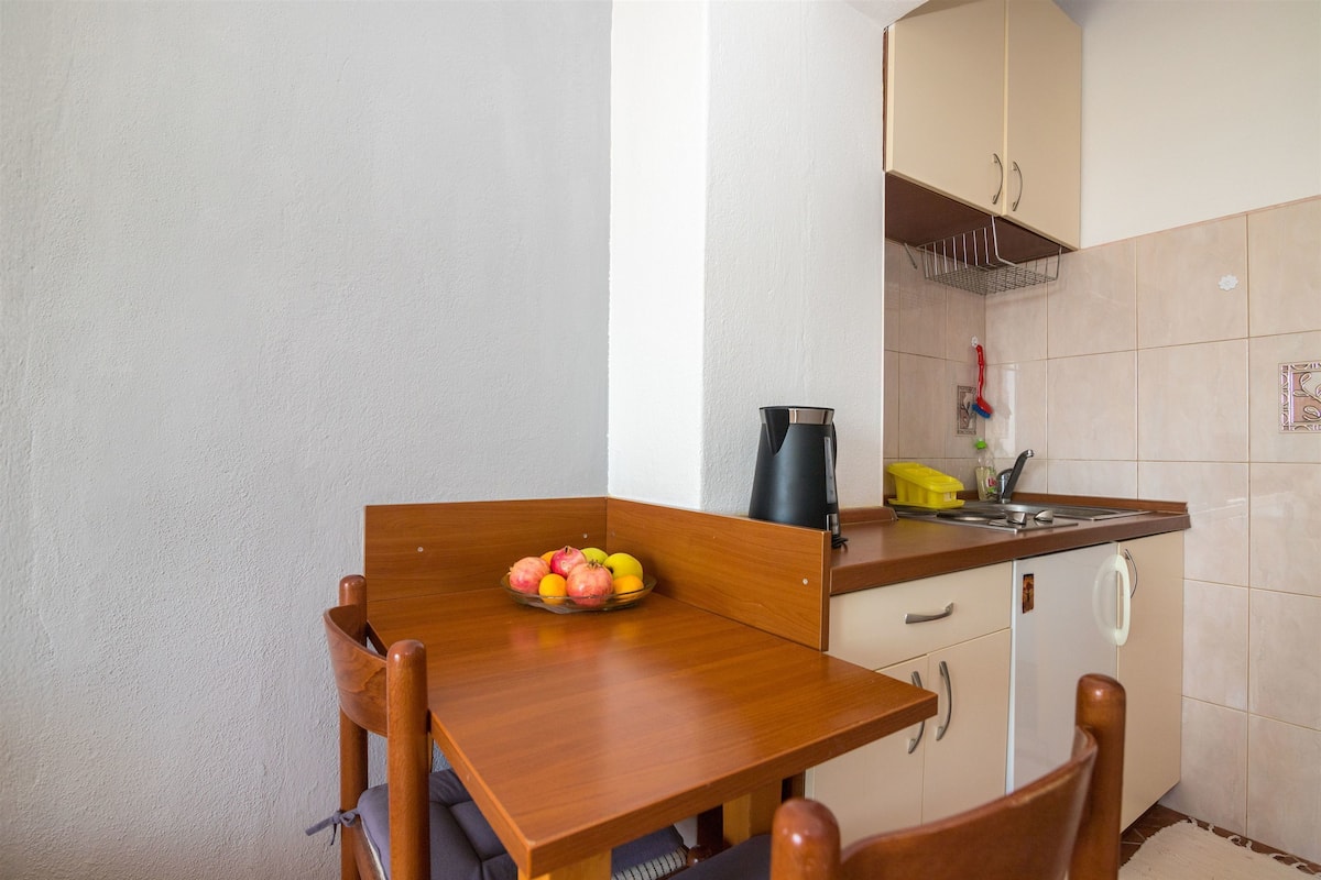 A small kitchen with a wooden table, a fruit bowl, a kettle, and a sink.