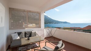 Un balcón con una mesa y sillas, una ventana y vistas al mar y a las montañas.