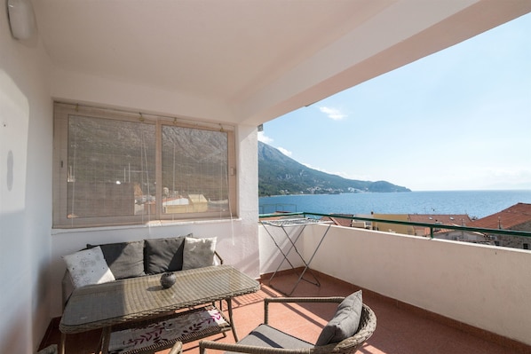 A balcony with a table and chairs, a window, and a view of the sea and mountains.