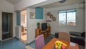 Una sala de estar con una mesa de comedor de madera, un sofá, un televisor sobre un mueble de madera y una ventana con vista al mar.