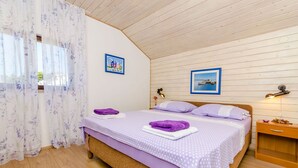 A bedroom with a wooden ceiling, a bed with purple bedding, two framed pictures on the wall, and a bedside table with a lamp. - Apartment Natasa (Okrug Gornji)
