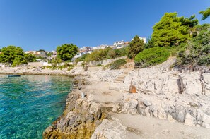 A coastal area with clear blue water, rocky terrain, and buildings in the background.