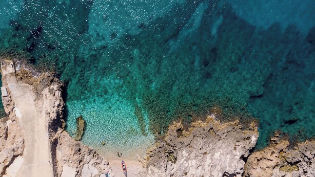 Una zona costera rocosa con aguas turquesas cristalinas, un sendero estrecho que lleva a una playa y un pequeño bote cerca de la orilla.