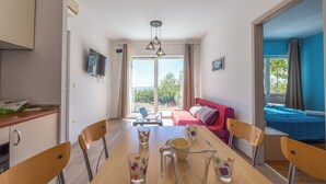 Una sala de estar moderna con una mesa de comedor, sillas y un sofá. Hay un televisor montado en la pared, una ventana con cortinas y vistas al exterior.