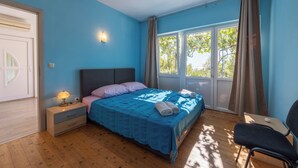 Un dormitorio con cama, mesita de noche, lámpara, silla y ventanas grandes con cortinas.