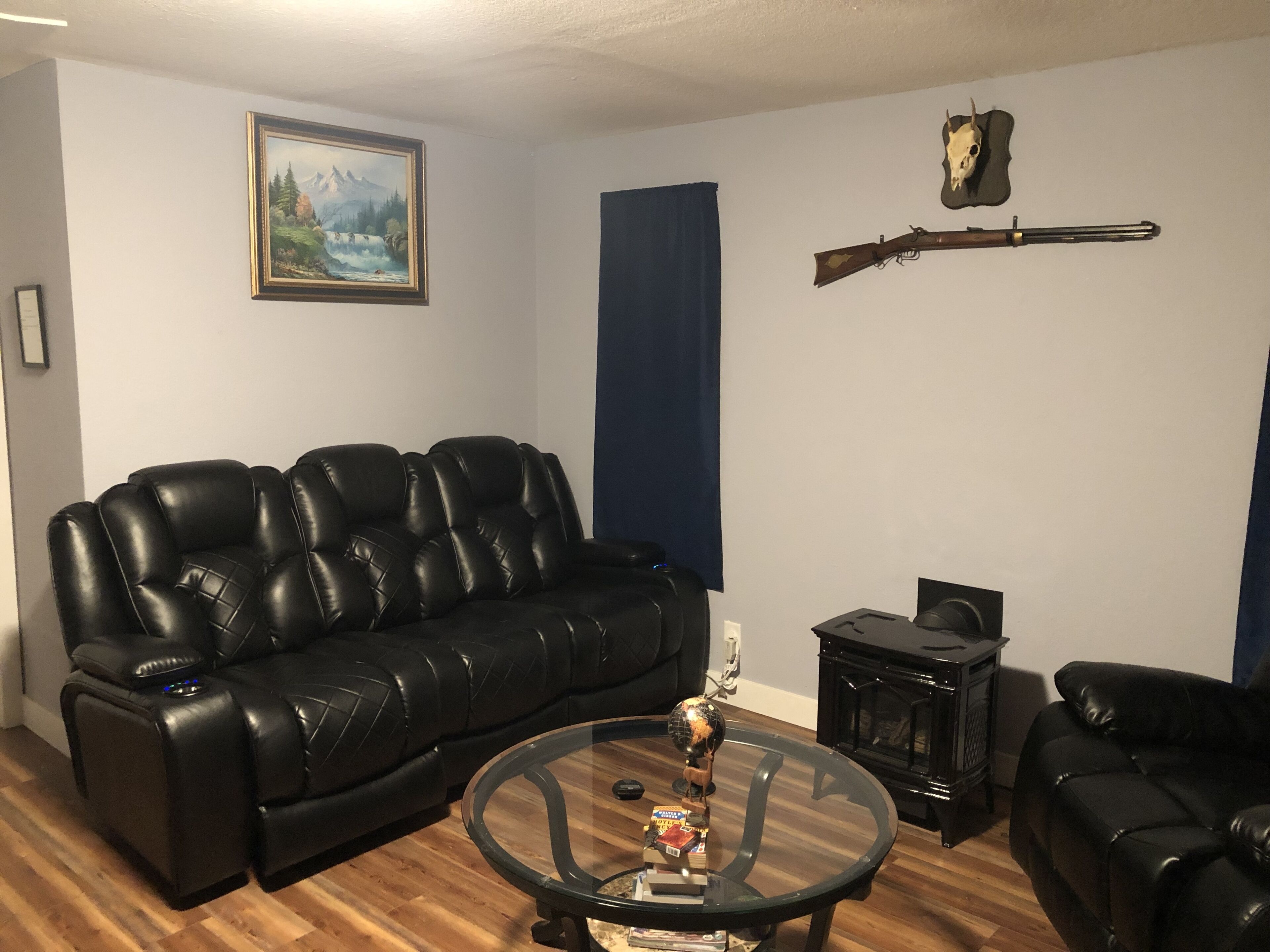 A living room with a black leather sofa, a glass coffee table with a globe, a fireplace, and a mounted rifle.