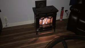 A small black wood stove with a visible fire, a chair, and a fire extinguisher in a room with wooden flooring and a white wall. - Fully private bunkhouse with all amenities. (Ashford)