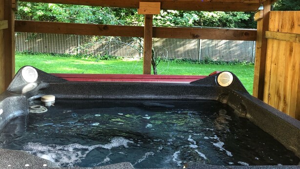 A hot tub under a wooden pergola with string lights, surrounded by a wooden fence and greenery. - Fully private bunkhouse with all amenities. (Ashford)