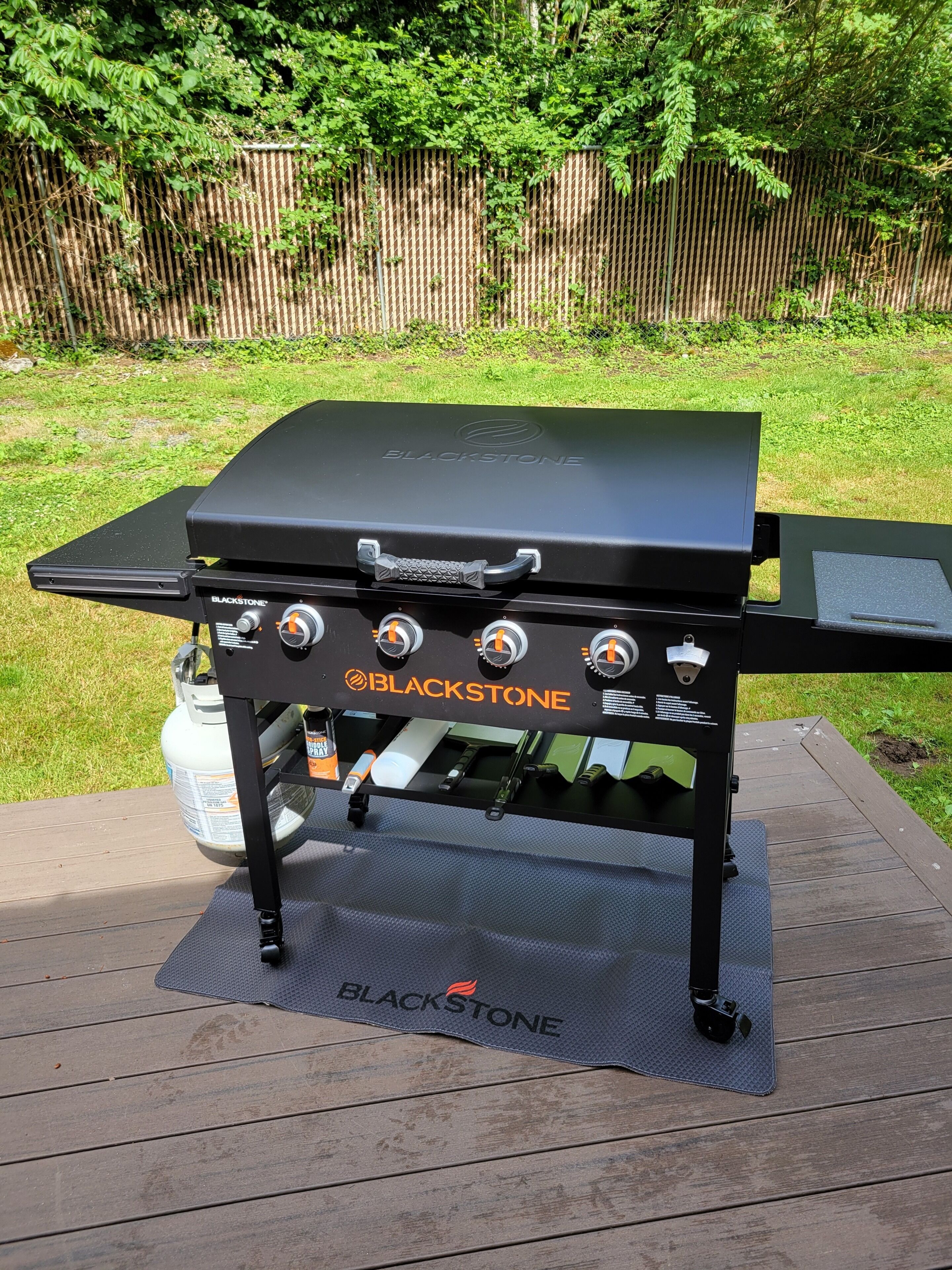 A BLACKSTONE grill with four burners, a side shelf, and a gas tank.