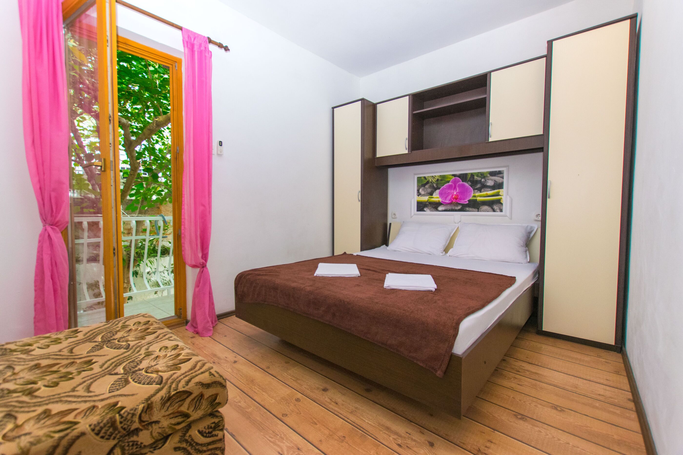 Une chambre à coucher avec un lit, une petite table, une fenêtre avec un tableau, et une porte coulissante avec des rideaux roses.