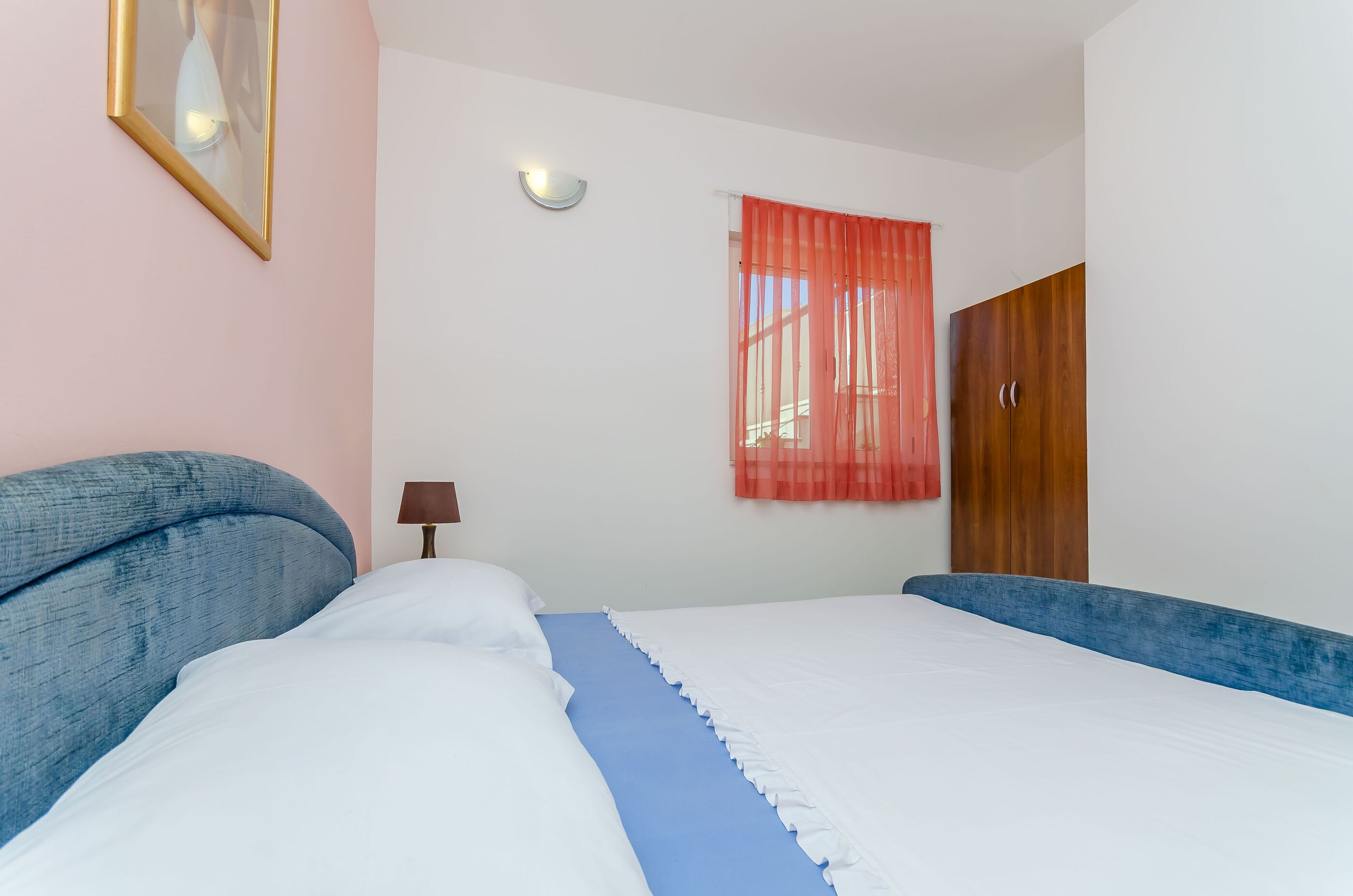 Een opgemaakt bed met wit en blauw beddengoed, een bedlampje, een ingelijst foto op de muur en een houten garderobe.