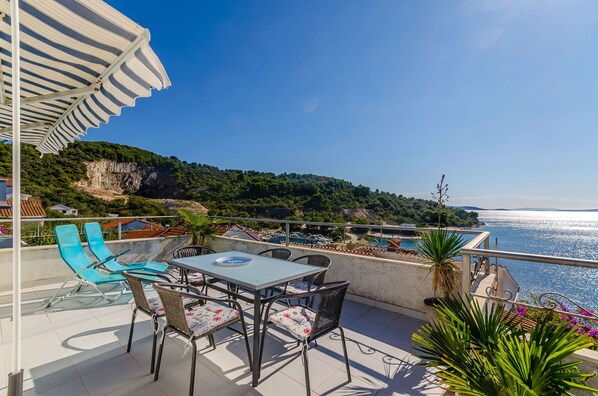 A terrace with a table and chairs, overlooking a coastal view with a pool and greenery.