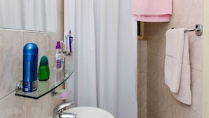 Un baño con un lavabo blanco, un estante de vidrio con artículos de aseo, una cortina de ducha y un perchero.