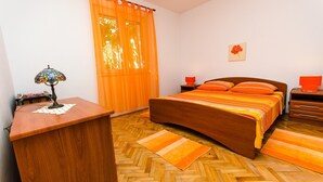 A bedroom with a wooden bed, orange curtains, a desk with a lamp, and a wooden floor. - Apartments Ivana (Trogir)