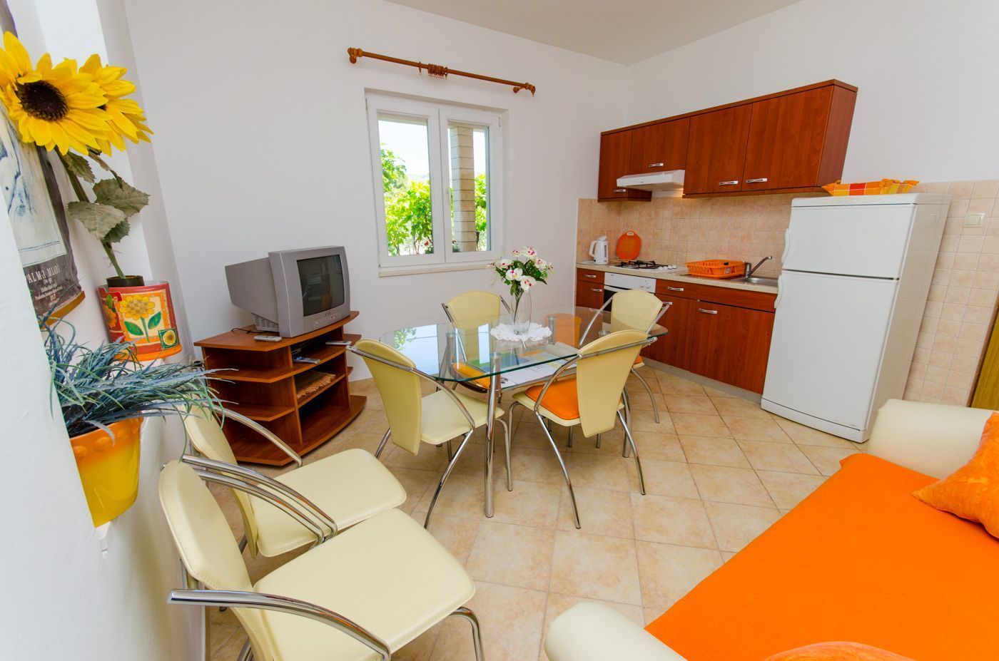 A compact kitchen with a refrigerator, stove, and sink. There is a dining table with chairs and a TV on a wooden stand.