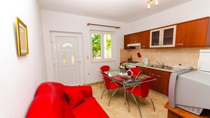 A compact living space with a red sofa, a glass dining table with red chairs, a kitchenette with wooden cabinets, and a white refrigerator. - Apartments Ivana (Trogir)