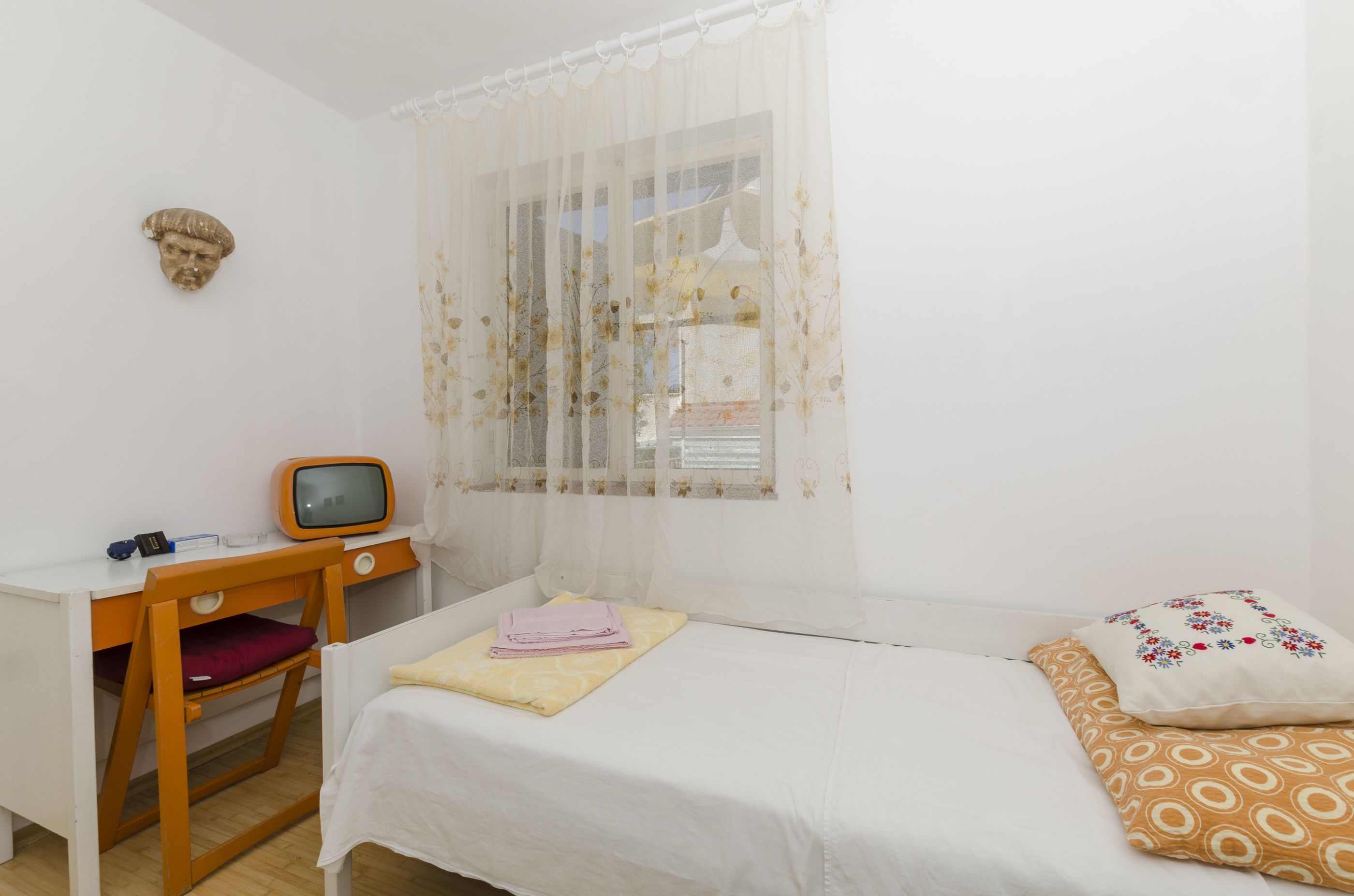 A bedroom with a bed, a desk with a television, and a window with sheer curtains.