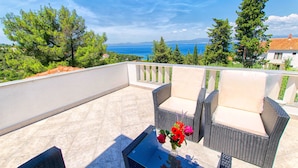 A balcony with wicker furniture and a glass table, overlooking a scenic view of the sea and greenery. - Apartments Damir - Katarina (Sutivan)