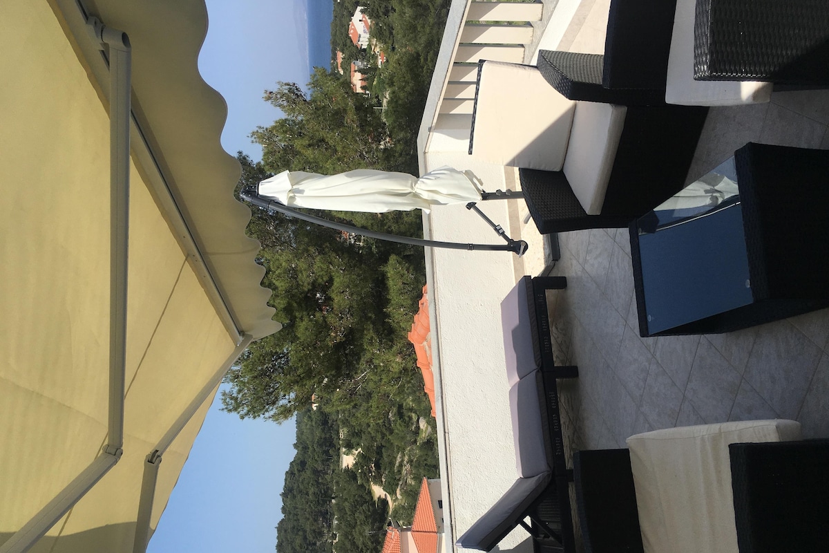 Eine Terrasse mit Sitzgelegenheiten im Freien, einem weißen Sonnenschirm und Blick auf eine Hügelkuppe mit Gebäuden.