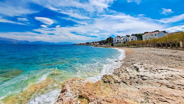 A coastal beach with clear turquoise waters, rocky shoreline, and a row of white buildings with balconies. - Apartments Damir - Katarina (Sutivan)