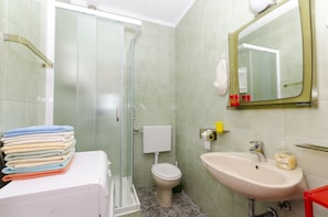 A bathroom with a glass shower door, a toilet, a sink with a mirror, and a stack of towels. - Apartments Simon (Bol)