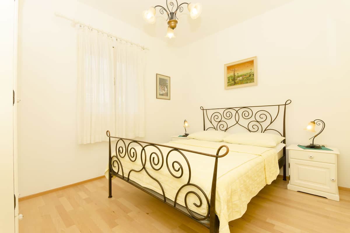 Un dormitorio con cama, mesita de noche, cuadro en la pared y ventana con cortinas.