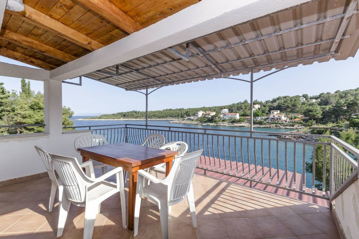 A covered outdoor seating area with a table and chairs, overlooking a body of water and a hillside with buildings.