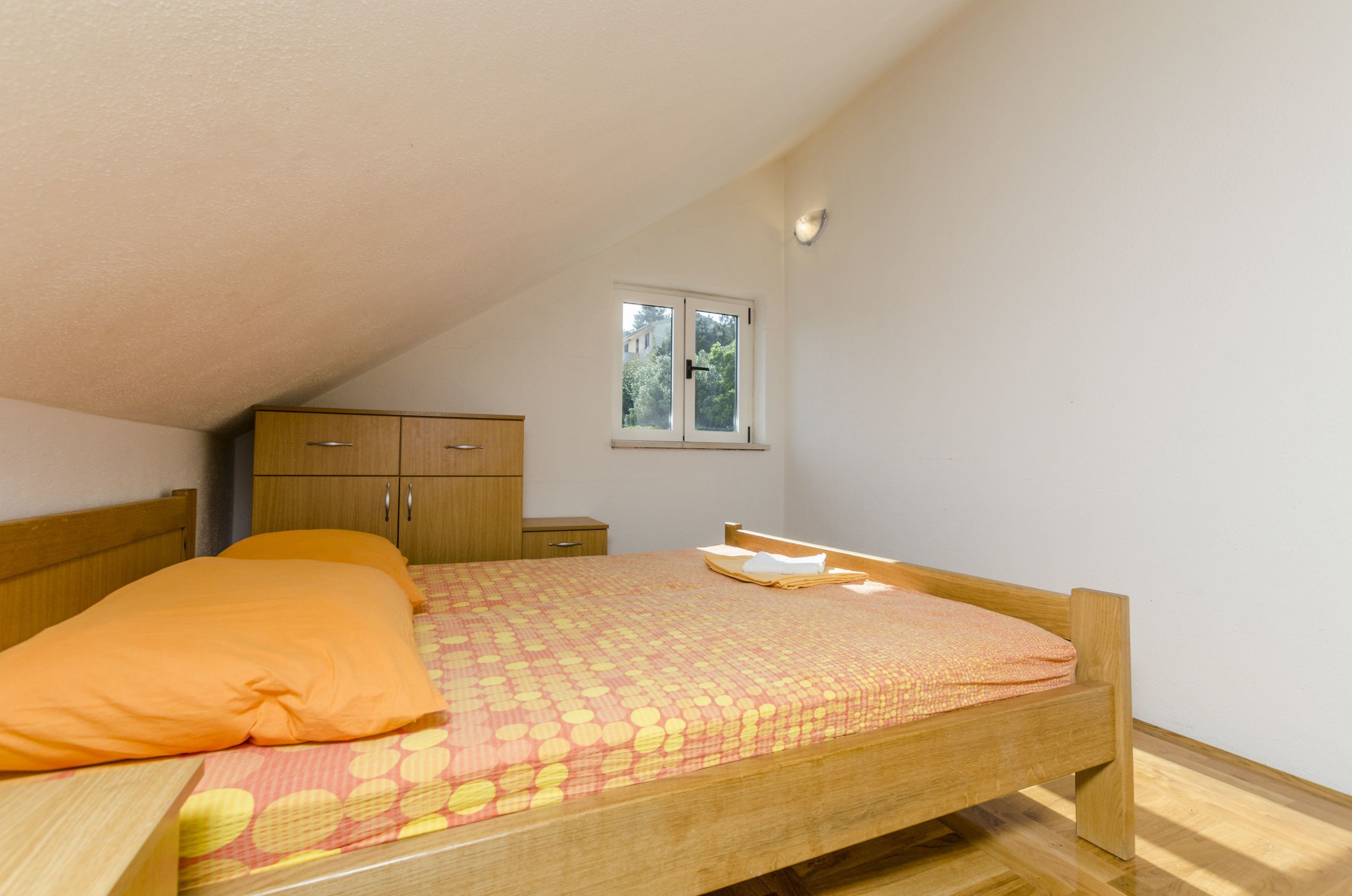 A bedroom with a sloped ceiling, a wooden bed, a nightstand, a wardrobe, and a window with a view of trees.