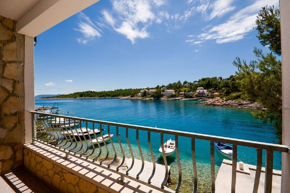 A balcony with a view of a calm sea, boats docked, and a stone wall.