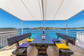 A covered outdoor seating area with a view of the sea and a distant town. - Apartment Mirjana (Rogoznica)