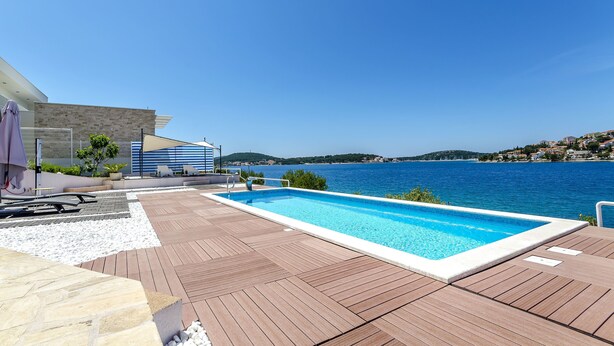 A swimming pool with a view of the sea, wooden deck, and a building with a blue and white facade. - Apartment Mirjana (Rogoznica)