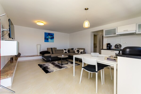 A modern living room with a sofa, coffee table, and dining area. There is a TV mounted on the wall, a ceiling light, and a kitchen area with a microwave and coffee maker. - Apartment Mirjana (Rogoznica)