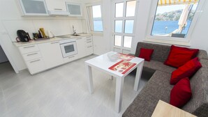A modern kitchen with white cabinetry, a dining table with a red placemat, and a grey sofa with red cushions. - Apartment Mirjana (Rogoznica)