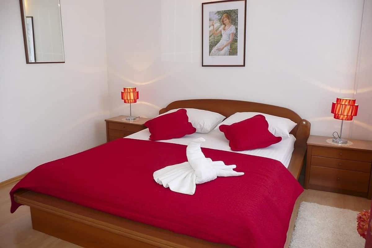 Ein ordentlich bezogenes Bett mit einer roten und weißen Tagesdecke, zwei roten Kissen und einem weißen Handtuch. Zwei Nachttische mit Lampen, ein gerahmtes Bild an der Wand und ein Spiegel über dem Bett sind zu sehen.