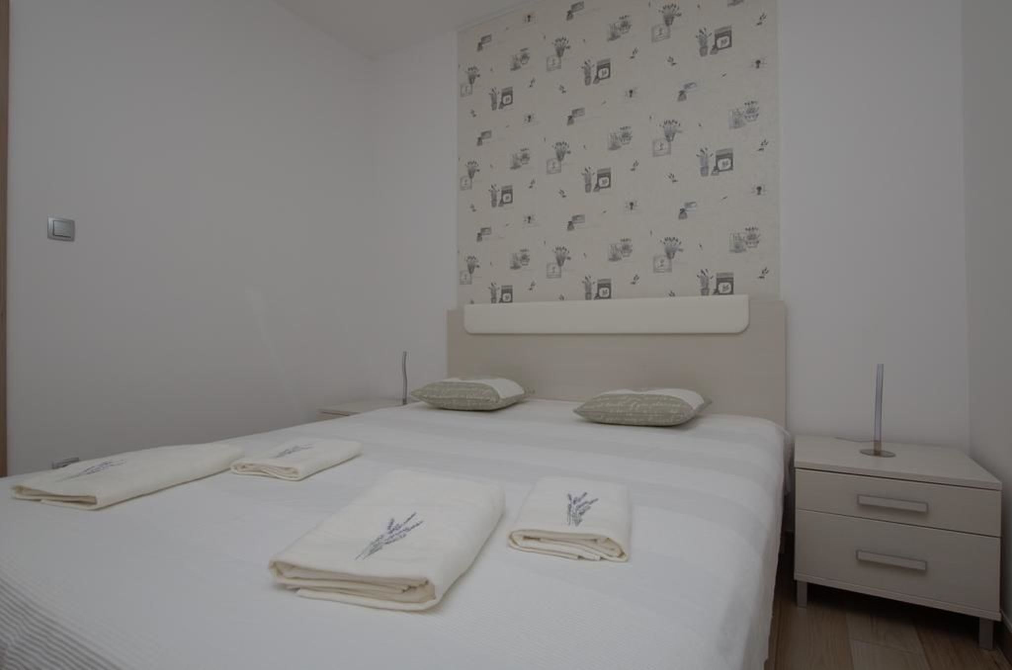 A hotel room with a neatly made bed, two pillows, a bedside table with a lamp, and a wall with a patterned wallpaper.