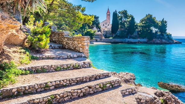 A coastal scene with stone steps leading to a building with a red tower, surrounded by trees and water. - Apartments Fani (Bol)