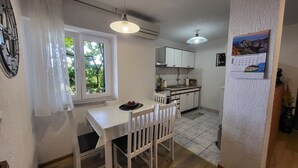 A compact kitchen with white cabinets, a dining table, and chairs. There is a window with curtains, an air conditioning unit, and a calendar on the wall.