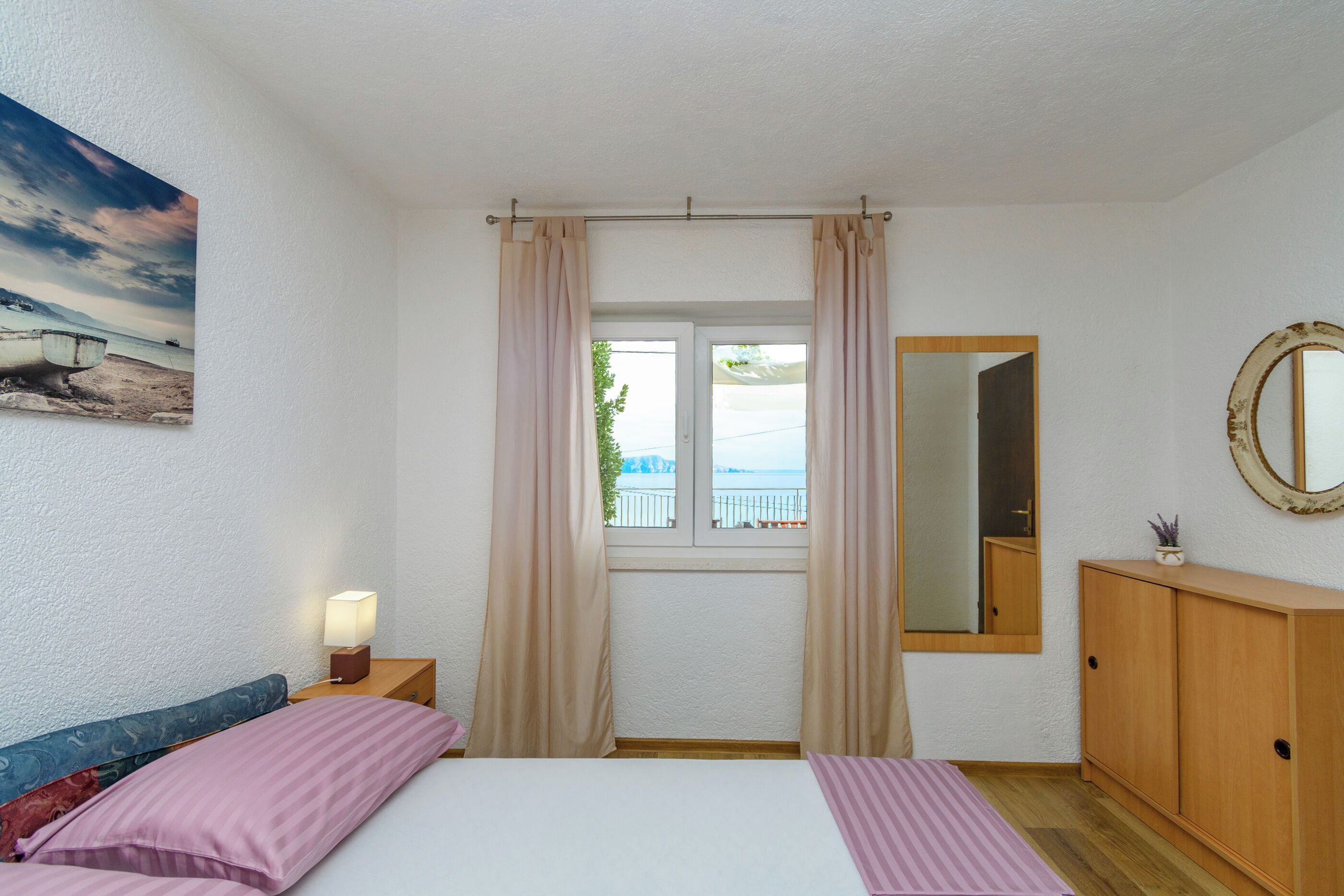 A bedroom with a bed, a nightstand, a lamp, a window with curtains, a mirror, and a view of the sea.