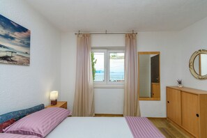 A bedroom with a bed, a nightstand, a lamp, a window with curtains, a mirror, and a view of the sea.